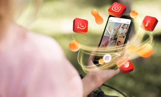 A person holding a smartphone displays a social media feed, surrounded by floating, vibrant icons of apps like Instagram and WhatsApp, set against a blurred green background, suggesting connectivity and interaction.