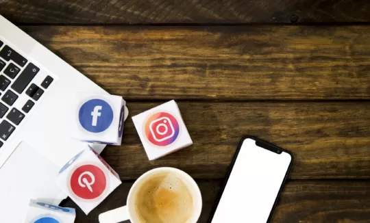 Wooden table with social media logo cubes, a cup of coffee, and a smartphone with a blank screen. The scene conveys connectivity and activity.